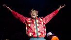 Jimmy Cliff hebt beide Arme in die Luft. Er trägt ein ausgefallen rot-kariertes Hemd und trägt eine Mütze dazu