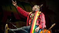 Jimmy Cliff sitzt auf einem Stuhl und erhebt die rechte Hand um das Publikum zu motivieren