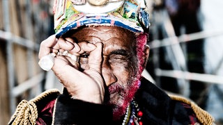 Der Jamaikaner Lee „Scratch“ Perry formt ein Loch mit den Fingern und hält es vor sein Auge. Er trägt grosse silberne Ringe an der rechten Hand