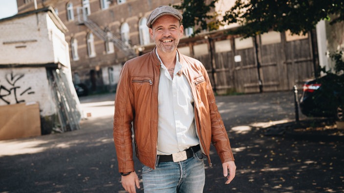 Das Bild zeigt den Arzt Doc-Esser mit grauer Schirmmütze - in weißem Hemd, Lederjacke und Jeans - gekleidet in einem Gebäudehof.