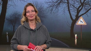 Yvonne Willicks steht im Servicezeit-Studio. Im Hintergrund ist das Bild einer dunklen Landstraße zu sehen. 