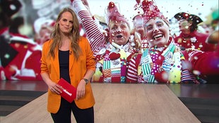 Svenja Kellershohn steht im Servicezeit-Studio. Im Hintergrund sieht man zwei verkleidete Menschen, die Karneval zelebrieren.