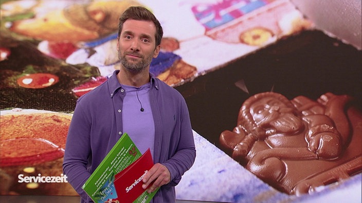 Der Moderator steht im Studio. Im Hintergrund ist ein Adventskalender zu sehen.