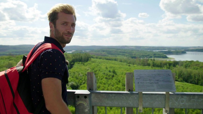 Daniel Aßmann steht an einem Geländer hoch oben über der Wald-Seen-Landschaft des Sauerlandes