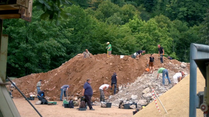 Mehrere Personen mit Schaufeln und Eimern auf einem braunen Erdhügel