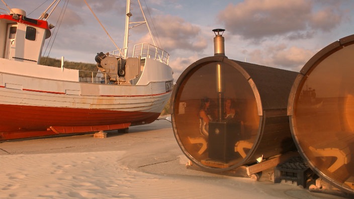 Zwei Fasssaunen stehen am Strand neben einem aufgedocktem Kutter