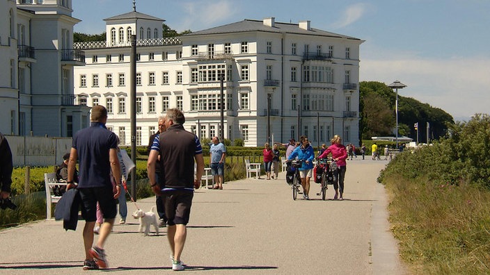 Andrea und Elke schieben  Fahrräder auf einer Promenade, im Hintergrund ein großes, weißes Gebäude