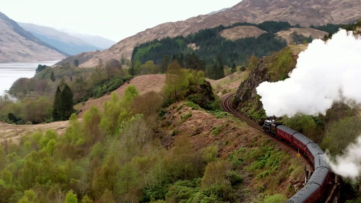 Ein mit einer Dampflokomotive gezogener Zug auf kurviger Streecke inmitten einer Berglandschaft