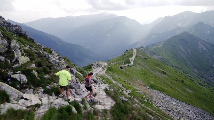 Magda und Ramon springen über Felsblöcke auf einem schmalen Bergpfad