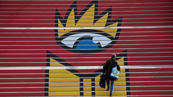 Eine Person steigt in der Glashalle der Neuen Messe in Leipzig eine Treppe mit dem Logo der Leipziger Buchmesse hinauf.