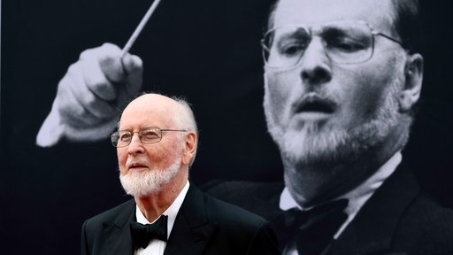 John Williams steht vor einem Plakat mit einem älteren Foto von ihm.