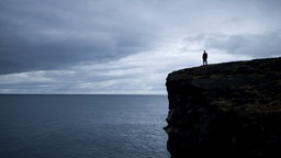 In der Ferne ist ein Mann zu sehen, der auf einem großen schwarzen Felsen steht und auf das endlose Meer hinaus blickt.