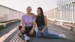 Die Hosts Helena und Melis sitzen auf einer Brücke und schauen in die Kamera