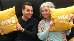 Ali Güngörmüs und Heike Knispel halten orangefarbene WDR 4-Kissen in die Kamera