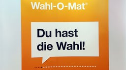 Zwei junge Leute stehen vor einen Bildschirm mit der Aufschrift "Du hast die Wahl! - Wahl-O-Mat".