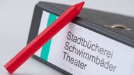 Roter Stift neben einem Aktenordner mit der Aufschrift "Stadtbücherei, Schwimmbäder, Theater"