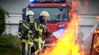 Zwei Feuerwehrmänner, die Atemmasken tragen löschen ein Feuer. Im Hintergrund steht ein Feuerwehrwagen.