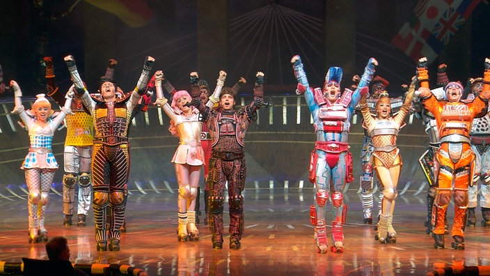 Das Ensemble des "Starlight Express" steht auf der Bühne singend und sie halten die Hände hoch.