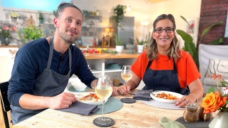 Moderatorin Yvonne Willicks und Profikoch Lars Middendorf sitzen am Tisch, vor ihnen stehen Gläser mit Wein und Teller mit Pasta.