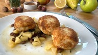 Das Gericht "Topfenknödel mit Apfel-Thymian-Kompott" auf einem weißen Teller, drumherum liegen Nüsse, Zitronen und Birnen.