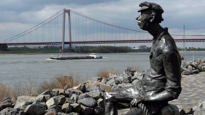 Die Bronzefigur Poortekeerl an der Emmericher Rheinpromenade, im Hintergrund die Hängebrücke