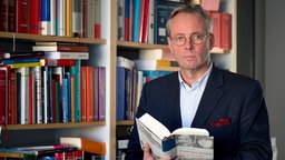 Butz Peters, Rechtsanwalt und RAF-Experte, steht vor einem Bücherregal und hält ein Buch in der Hand.