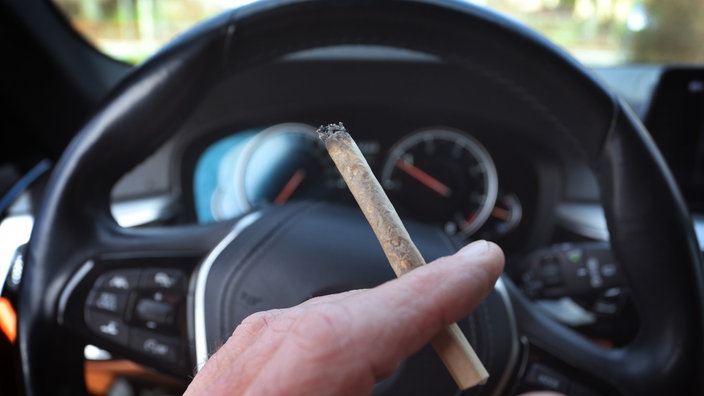 Ein Mann sitzt mit einem Joint zwischen den Fingern am Steuer eines Autos