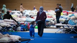 Eine Frau läuft Hand in Hand mit einem Kind durch eine Turnhalle. Es sind viele Feldbetten errichtet, auf denen teilweise persönliche Gegenstände und Klamotten liegen.