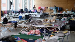 In einer Turnhalle sind Feldbetten aufgestellt. Darauf befinden sich teils persönliche Gegenstände und Klamotten. Im Hintergrund sind Menschen zu verzeichnen.