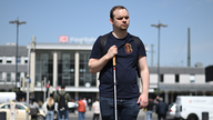 WDR-Reporter Kevin Barth steht mit Langstock in der Hand vor dem Dortmunder Hauptbahnhof