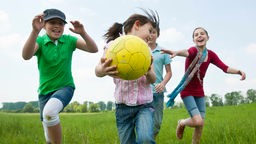Kinder rennen mit Ball über eine grüne Wiese 