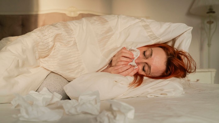 Eine kranke Frau liegt auf dem Bett und reibt sich die Nase mit einem Papiertaschentuch.