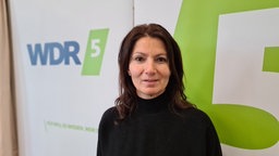 Frau mit schwarzen Haaren steht vor einem WDR 5 Logo