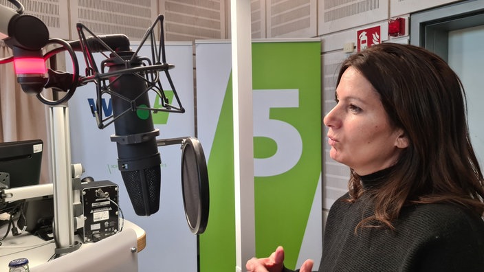 Eine Frau mit langen, dunklen Haaren steht in einem Hörfunkstudio vor einem Mikrofon