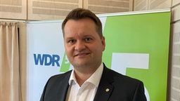 Ein Mann im Anzug mit weißem Hemd und kurzen braunen Haaren steht im WDR-5-Sendestudio vor einem Aufsteller mit dem Logo von WDR 5