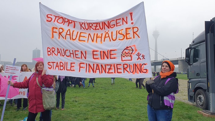 Zwei Frauen halten ein großes Transparent mit der Aufschrift "Stoppt Kürzungen! Frauenhäuser brauchen eine stabile Finanzierung!"