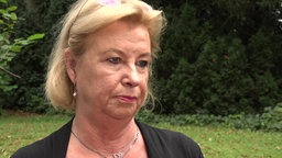 Ein Frau mit hellblonden Haaren und Perlenohrringen steht vor einer grünen Hecke und gibt ein Interview