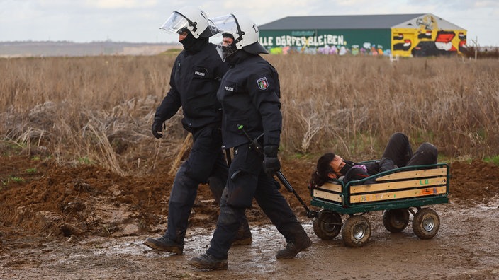 Zwei Polizisten räumen das besetzte Dorf Lützerath im Rheinischen Braunkohlerevier und ziehen einen Demonstranten in einem Bollerwagen hinter sich her