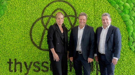 Vor einer grünen Wand mit Thyssenkrupp-Logo stehen NRW-Wirtschaftsministerin Mona Neubaur, Bundeswirtschaftsminister Robert Habeck und der Thyssenkrupp-Vorstandschef Miguel López