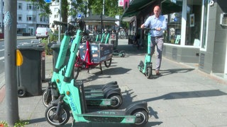 Im Vordergrund stehen vier E-Scooter aufgereiht, in Hintergrund schiebt ein Mann einen E-Scooter zum Parkplatz