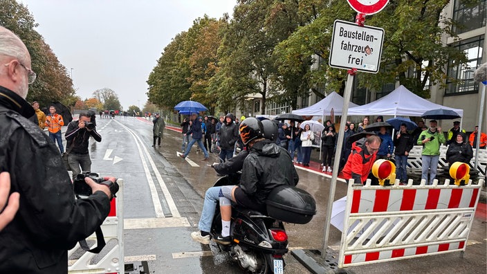 Mehrere Menschen sind auf offener Straße an einer Baustelle versammelt, in der Mitte fahren zwei Personen auf einem Motorrad.