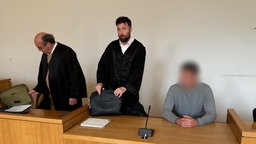 Auf dem Foto sind zwei Männer in typischer Juristenkleidung, daneben ein kurzhaariger Mann in einem grau-melierten Pullover.