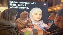 Frauen aus verschiedenen Kulturen stehen vor einem Plakat mit der Aufschrift "Männerberuf ? Bis jetzt vielleicht"