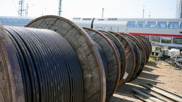 Dutzende große Kabeltrommeln liegen aufgereiht auf einer Gleisanlage. Im Hintergrund steht ein Intercity.