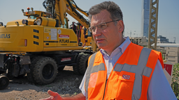 Ein Mann mit Brille, Hemd und orangener Warnweste steht vor einem gelben Bagger auf einer Gleisanlage.