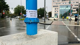 Ein Betonklotz auf dem ein blauer Metallpfeiler steht. Darauf klebt ein Plakat mit dem Text: Ist das Kunst oder kann das weg?