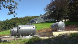 Halbfertige Heizungsanlage mit drei Heizkesseln auf einem Betonplateau auf einer Wiese. Dahinter ein großes lang gezogenes Gebäude aus Schiefer