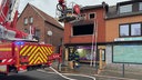 Das Bild zeigt ein Feuerwehrauto vor einer Häuserfront, in der Mitte ein Haus mit einem verrußten Fenster ohne Scheiben