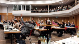 Mehrere Kreisabgeordnete sitzen an langen Holztischen im Kreishaus und heben die Hand.