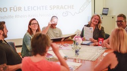 An einem runden Tisch sitzen mehrere Menschen, lachen, im Hintergrund eine große Leinwand